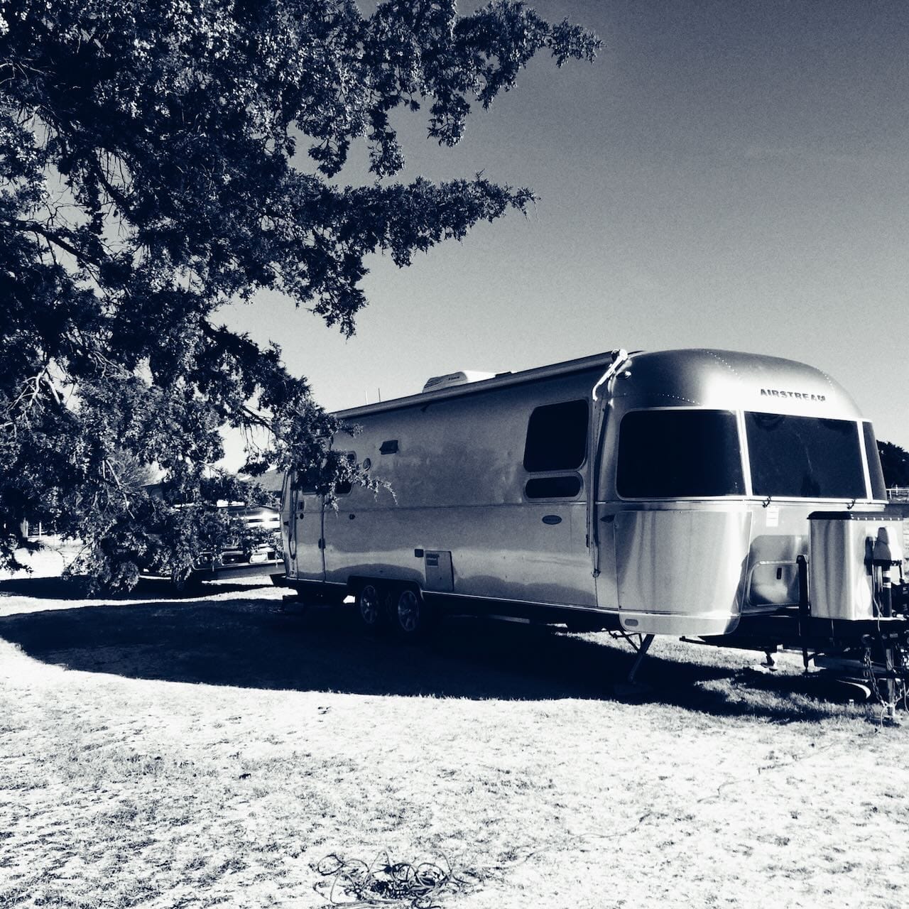 B&W photo of airstream in a fields, under trees with a pickup in the background.