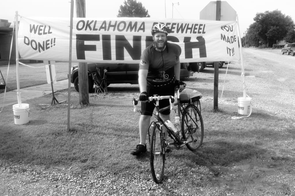 B&W picture of myself leaning against a bicycle at a banner marking the finish of Oklahoma Freewheel.