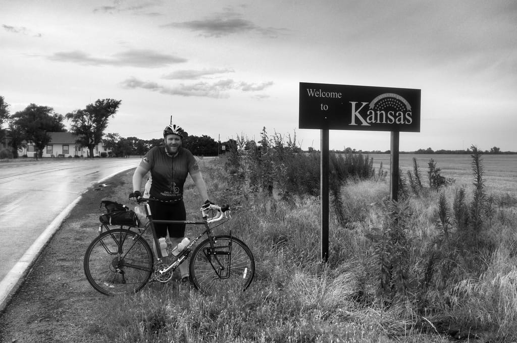B&W picture of myself leaning against a bicycle at a sign marking the Kansas border.
